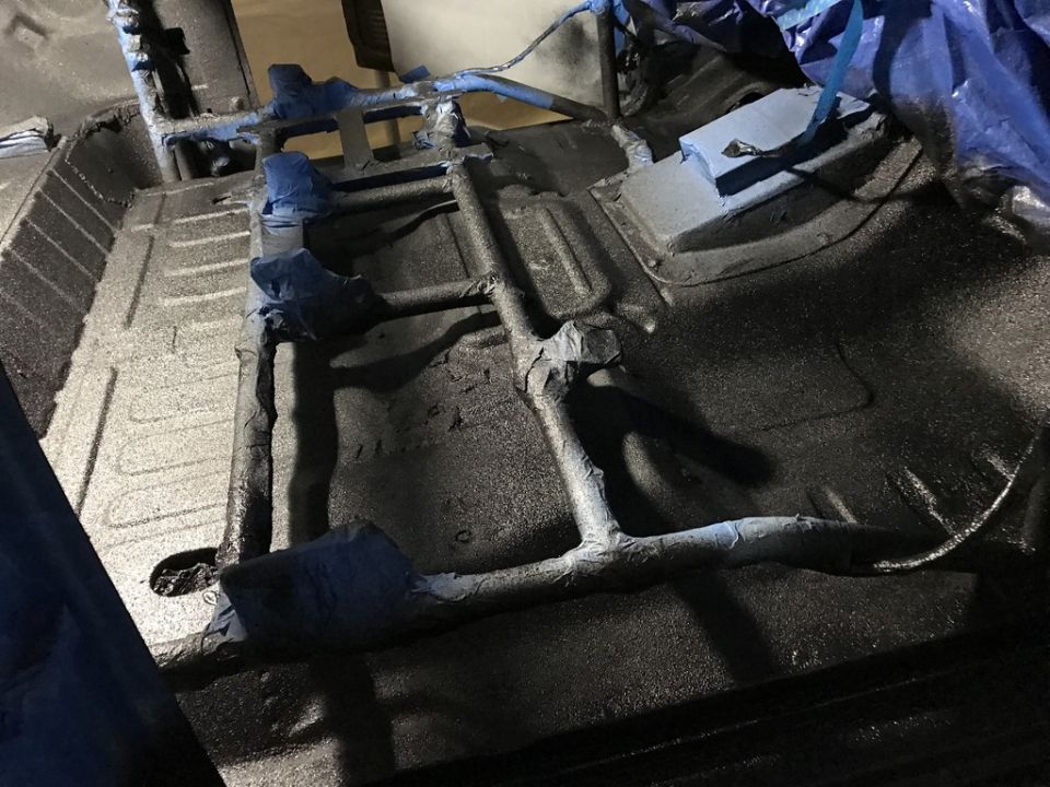 Vehicle undercarriage with frame rails and floor pan coated in black protective material, some parts masked with blue tape