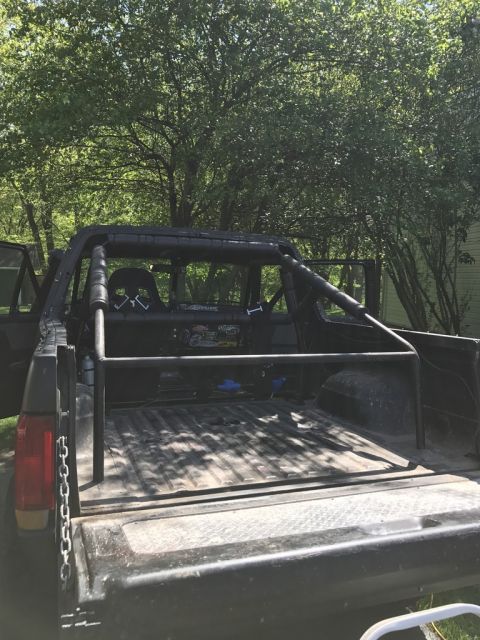 1989 Ford Bronco - Looking into a pickup bed fitted with a black tubular roll bar and rear chase rack, parked under trees.