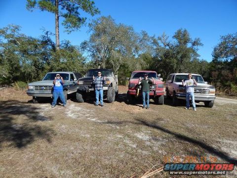 1989 Ford Bronco - Four trucks and SUVs are parked in a grassy area with trees in the background under a clear blue sky.
