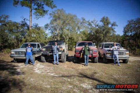 1989 Ford Bronco - Four lifted trucks and SUVs are parked in a dirt lot with people standing in front of them.