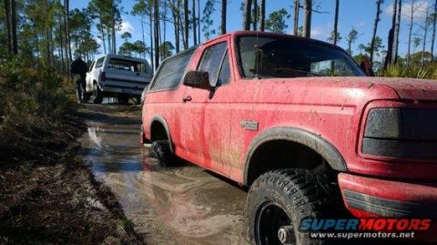 1989 Ford Bronco - Two Broncos navigate a muddy trail through a wooded area, with the red one in the foreground appearing stuck.