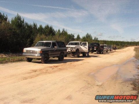 1989 Ford Bronco - A convoy of SUVs, including a 1987 Ford Bronco, travels down a dusty dirt road lined with trees.