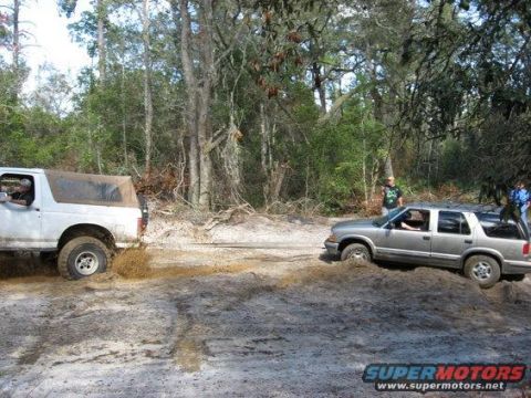1989 Ford Bronco - Two SUVs navigate a muddy trail in a wooded area, with one vehicle appearing to be stuck.