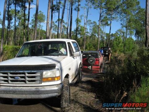 1989 Ford Bronco - A white 1989 Ford Bronco navigates a muddy off-road trail, followed by other vehicles in a wooded area.