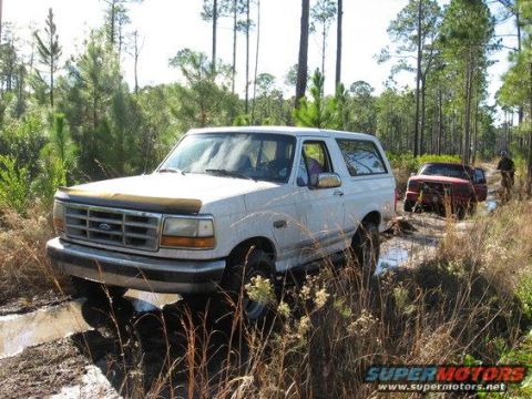 1989 Ford Bronco - White 1989 Ford Bronco navigates a muddy path in a wooded area, with other vehicles behind.