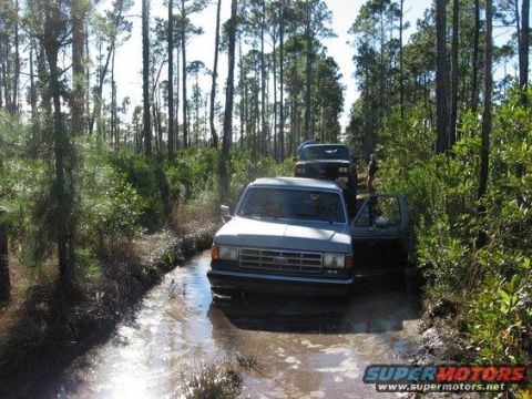 1989 Ford Bronco - Two trucks navigate a water-filled path in a wooded area.