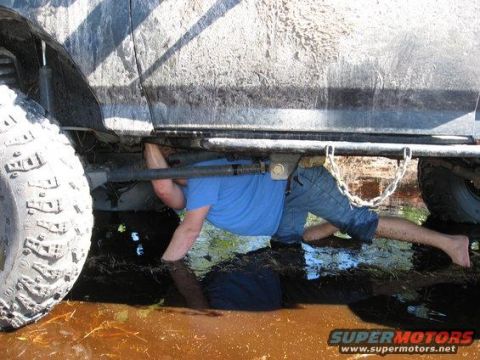 1989 Ford Bronco - A person works on the undercarriage of a dirty vehicle submerged in mud and water, with a chain hanging from the frame.