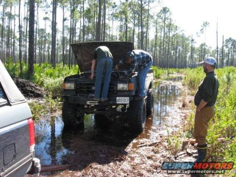 1989 Ford Bronco - Two men work on a 1989 Ford Bronco stuck in a muddy, wooded area while another watches.