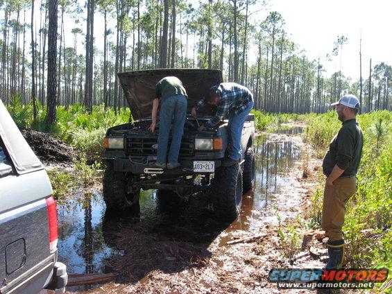 1989 Ford Bronco with hood open, stuck in mud, two people working on engine, another person watching