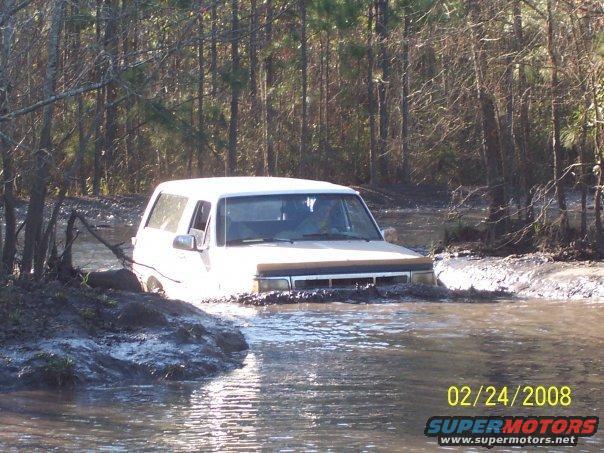 White 1989 Ford Bronco driving through muddy water, front 3/4 view
