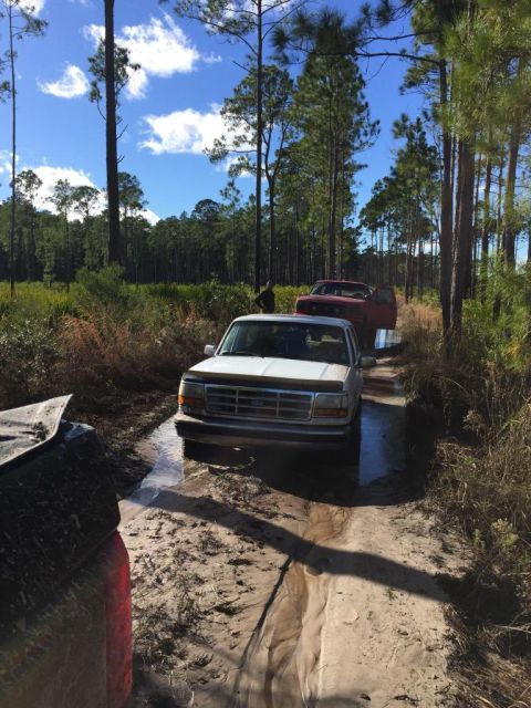 1989 Ford Bronco - A white Ford F-150 navigates a muddy trail, with another vehicle following behind.