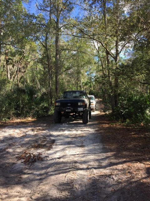 1989 Ford Bronco - Two trucks navigate a sandy path through a wooded area on a sunny day.