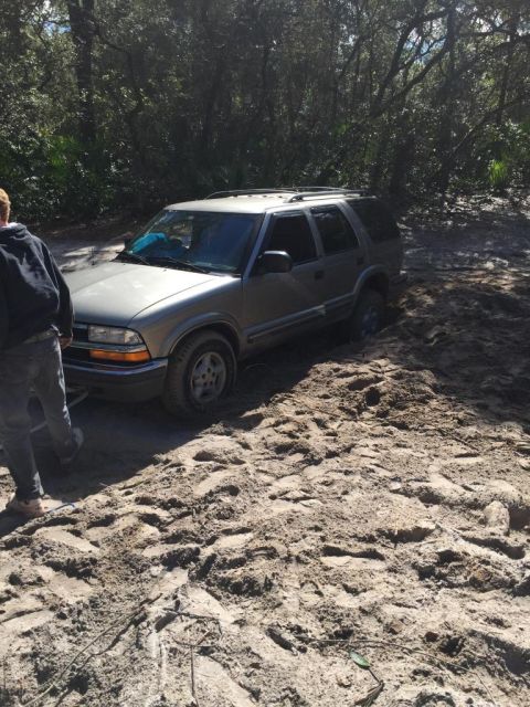 1989 Ford Bronco - A silver SUV, likely a Chevrolet Blazer from the late 1990s, is stuck in a sandy area with trees in the background.