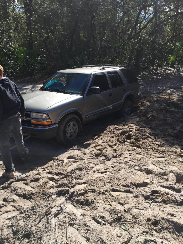 Silver SUV stuck in sand with a person standing nearby. The vehicle appears to be a Chevrolet Blazer from the late 1990s