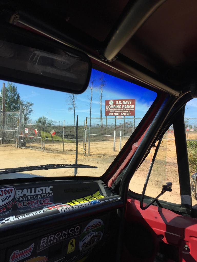 View from inside a vehicle looking out at a U.S. Navy Bombing Range sign and fence. Dashboard has many stickers, including one that says 'BRONCO GRAVEYARD.COM'