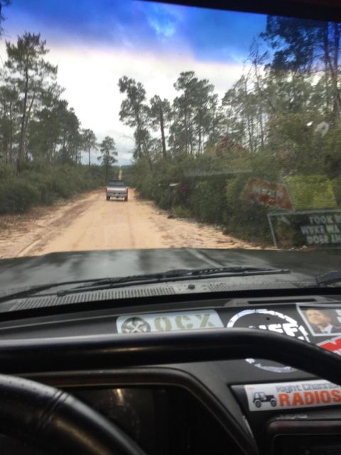1989 Ford Bronco - Two trucks drive down a dusty dirt road surrounded by trees under a cloudy sky.