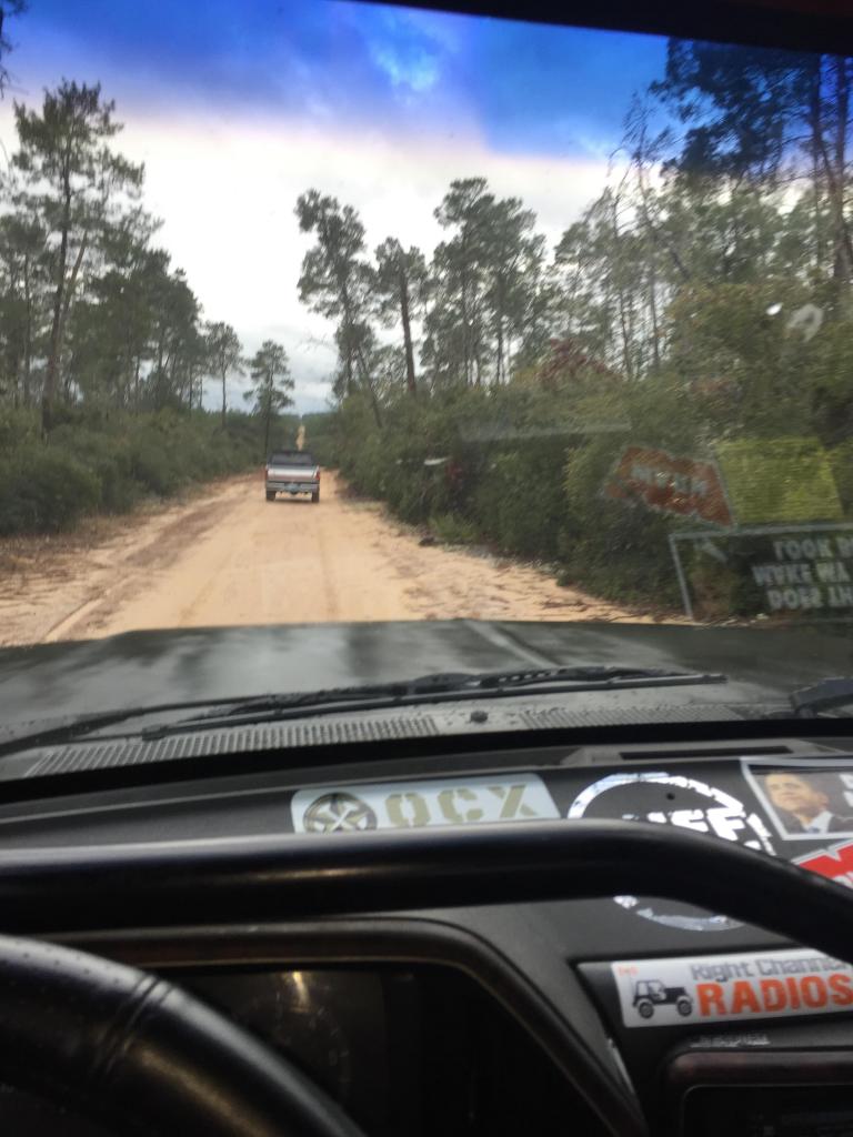 View from inside a vehicle looking down a dirt road at another truck ahead. Trees line both sides of the road
