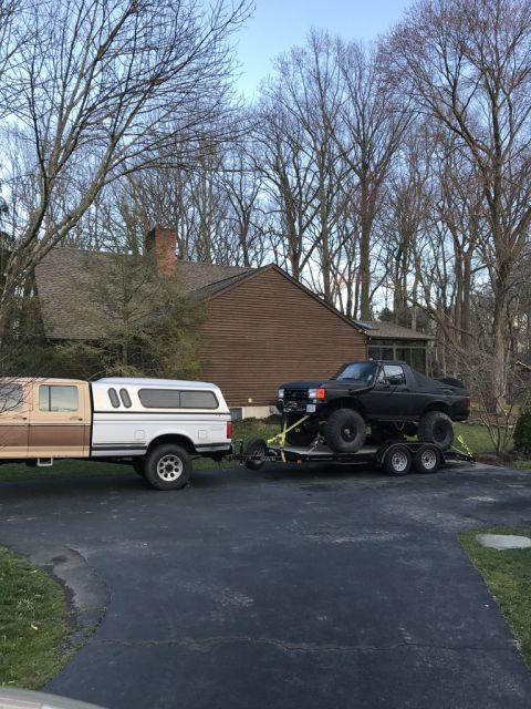 1989 Ford Bronco - A brown and white pickup truck tows a black lifted truck on a trailer in a driveway.
