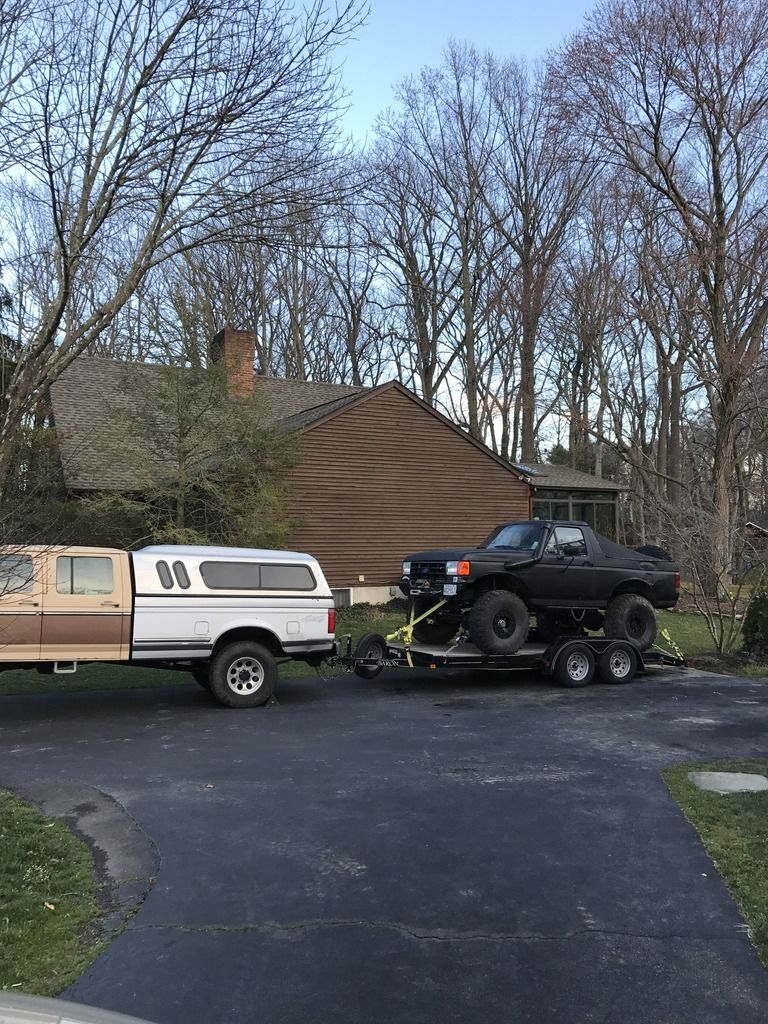 Brown and white pickup truck towing a black lifted truck on a trailer. The black truck appears to be a Ford Bronco