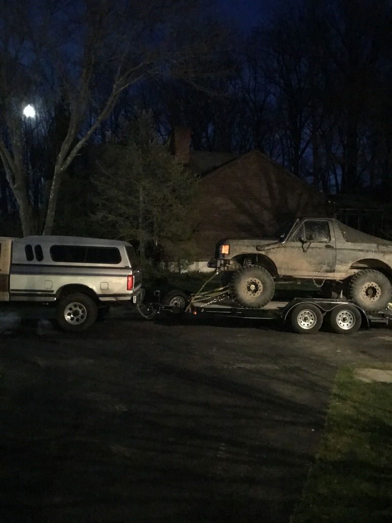 Mud-covered Ford Bronco on a trailer, towed by a pickup truck with a camper shell