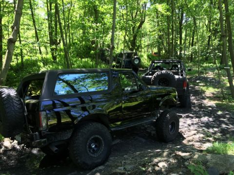 1989 Ford Bronco - A black 1989 Ford Bronco navigates a rocky, wooded trail with other off-road vehicles in the background.