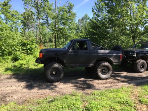 1989 Ford Bronco - A modified dark gray Ford Bronco is parked on a dirt path, with another truck visible behind it.