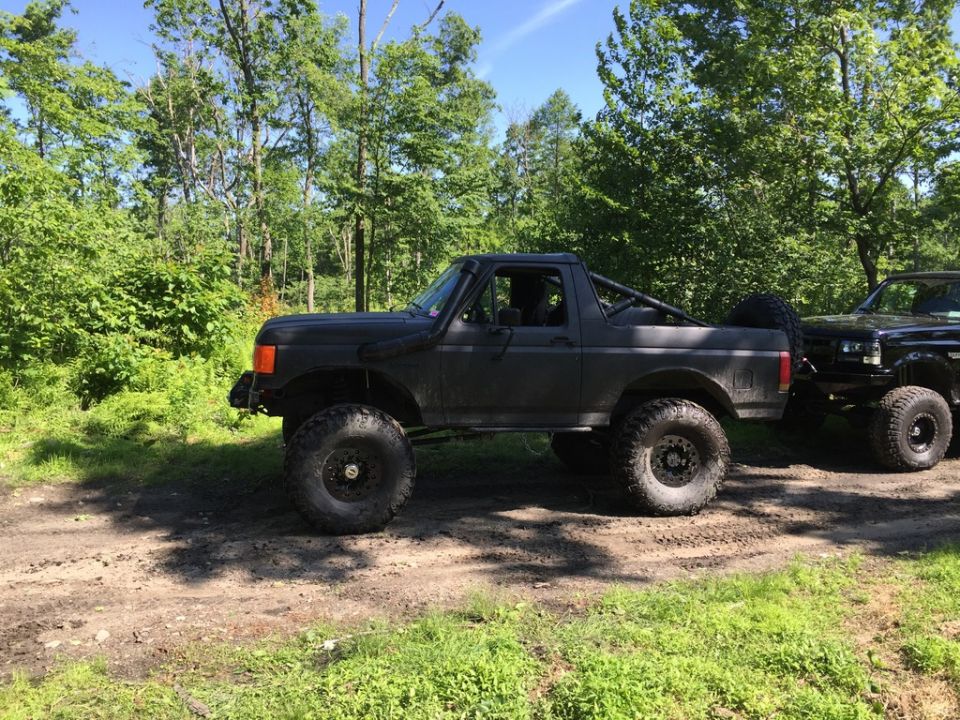 Dark gray Ford Bronco with large tires and a roll cage, parked on a dirt road next to another black truck