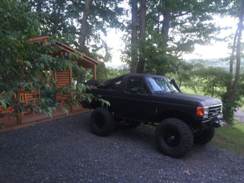 1989 Ford Bronco - Modified 1989 Ford Bronco sits parked on a gravel driveway near a cabin in a wooded area.