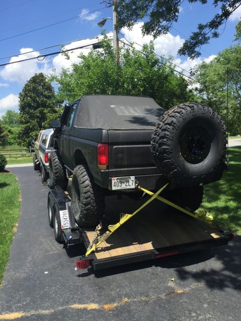 1989 Ford Bronco - A lifted black Bronco with a soft top and large spare tire is secured to a trailer, with another vehicle visible behind it.