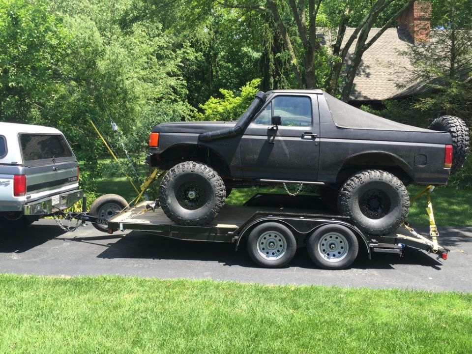 Black Ford Bronco on a dual-axle trailer, towed by a silver pickup truck. The Bronco has large off-road tires and a snorkel