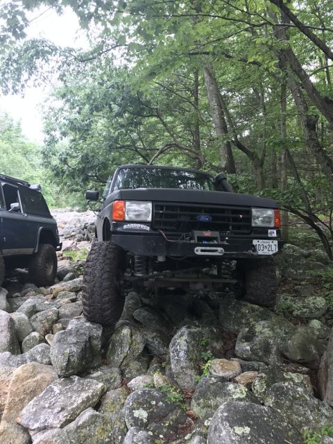 1989 Ford Bronco - A black 1989 Ford Bronco tackles a challenging rocky trail, with another off-road vehicle visible in the background.