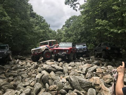 1989 Ford Bronco - Multiple off-road vehicles are seen navigating a challenging rocky path surrounded by trees.