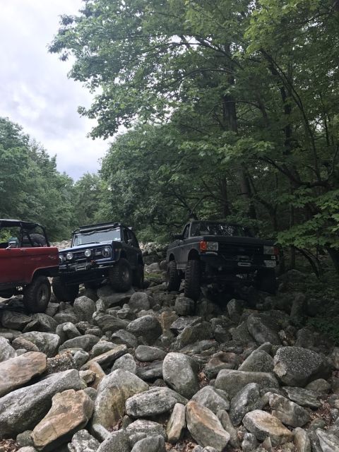 1989 Ford Bronco - Modified trucks traverse a challenging rocky trail surrounded by dense foliage.