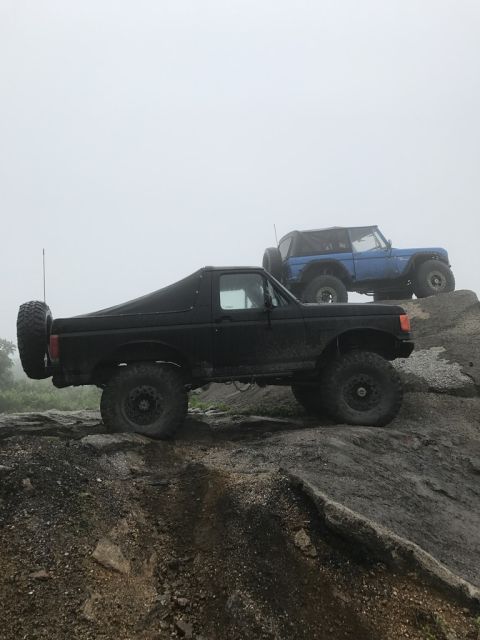 1989 Ford Bronco - Two off-road vehicles, a black Ford Bronco and a blue Jeep Wrangler, navigate a rocky incline on a foggy day.