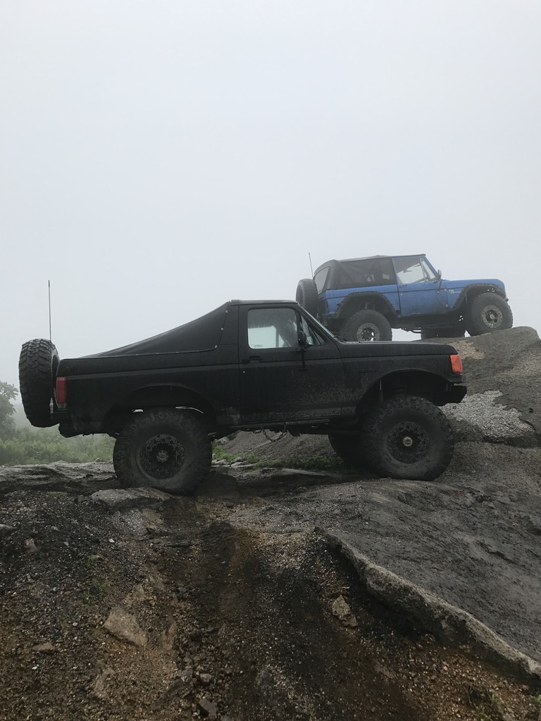 Black Ford Bronco with spare tire on rear, side view, on rocky terrain. Blue Jeep Wrangler behind