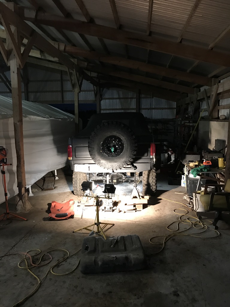 Gray Ford Bronco rear, lifted, with spare tire mounted. Underneath is being worked on in a dimly lit garage