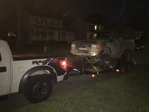 1989 Ford Bronco - A white Ford F-150 Raptor tows a mud-covered, dark-colored SUV on a trailer at night. The SUV appears to be a Ford Bronco.