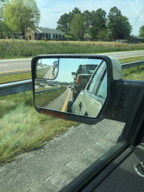 1989 Ford Bronco - The driver's side mirror reflects a line of vehicles on a highway, including a pickup truck towing a trailer. The reflection shows the side…