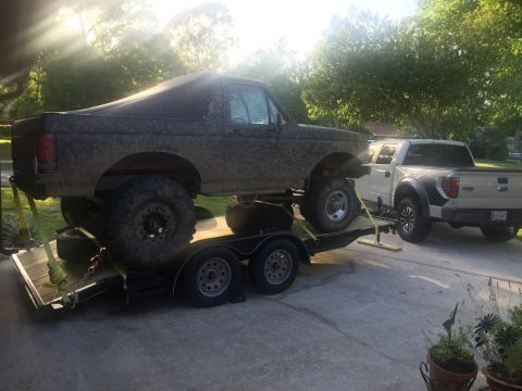 1989 Ford Bronco - A muddy, lifted 1980s Ford Bronco is loaded onto a flatbed trailer, with a white Ford F-150 Raptor parked behind it. The scene is set in a…