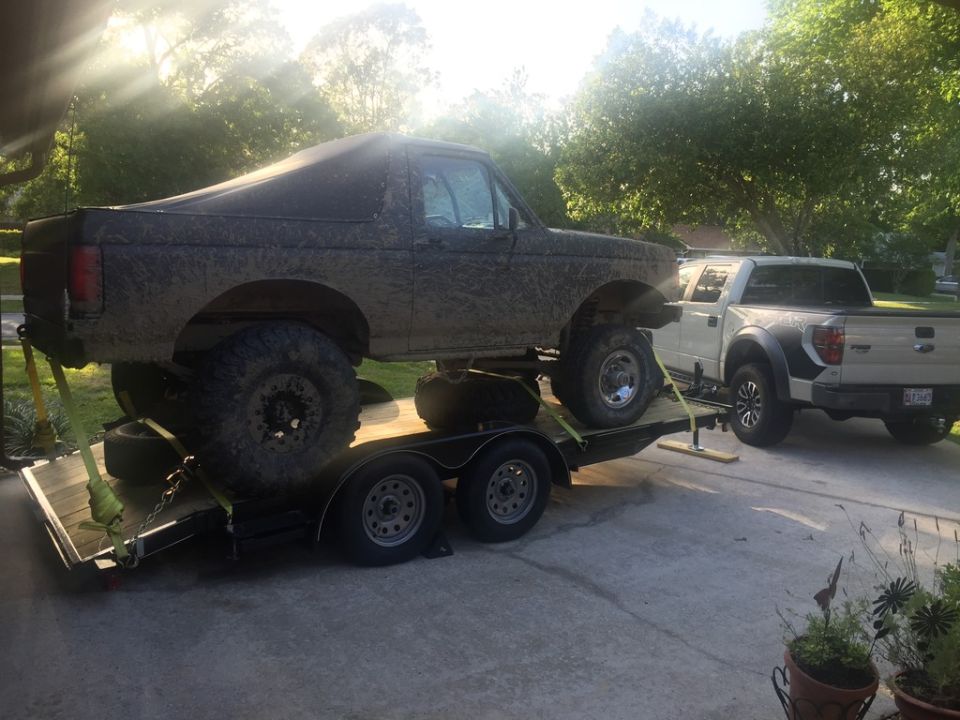Muddy 1980s Ford Bronco on a trailer, with a white Ford F-150 Raptor behind it.