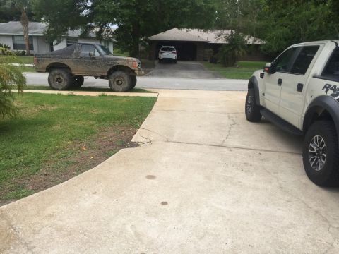 1989 Ford Bronco - A lifted, mud-covered 1989 Ford Bronco is parked on the street next to a driveway. A white Ford F-150 Raptor is parked in the driveway.