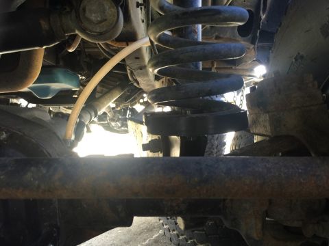 1989 Ford Bronco - Undercarriage view of a vehicle, showing a large coil spring, suspension components, and a portion of a tire. A yellow hose is routed near…