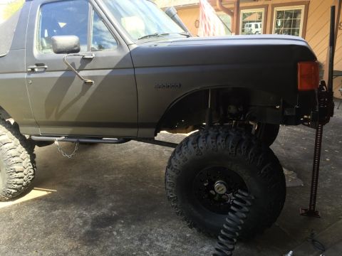 1989 Ford Bronco - The driver's side of a lifted, dark-colored Ford Bronco is shown, with a high-lift jack positioned next to the front tire. The vehicle feat…