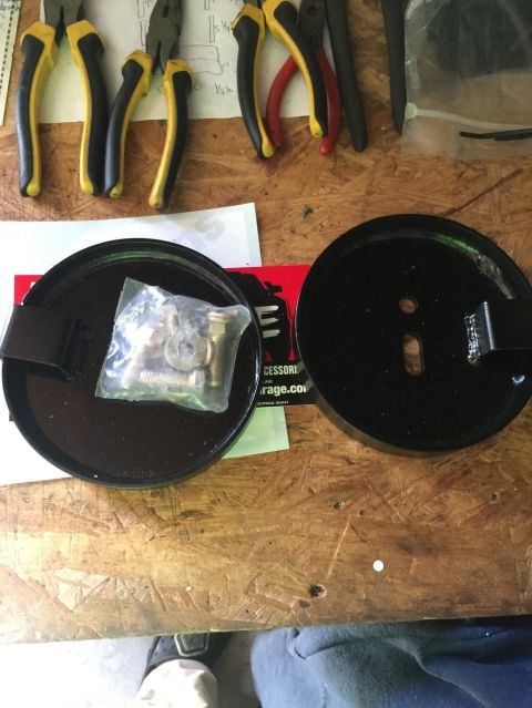 1989 Ford Bronco - Two black circular metal parts are laid out on a wooden surface. One part contains a clear plastic bag filled with bolts and washers.