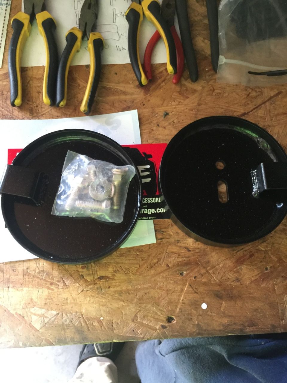 Two black circular metal parts, one with a bag of hardware, on a wooden surface.