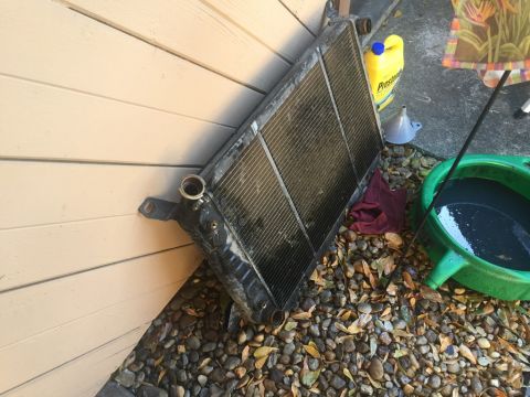 1989 Ford Bronco - A used car radiator is leaning against a beige wooden wall, surrounded by gravel and fallen leaves. A yellow jug of Prestone coolant, a fun…