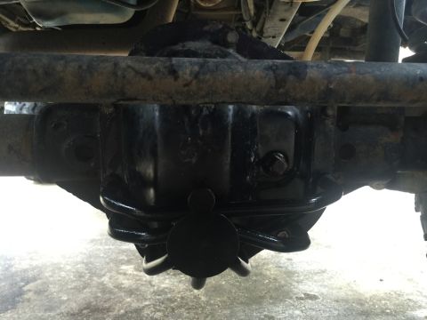 1989 Ford Bronco - The underside of a vehicle shows a black differential housing with a black cover and a black spring. A rusted metal bar runs horizontally a…