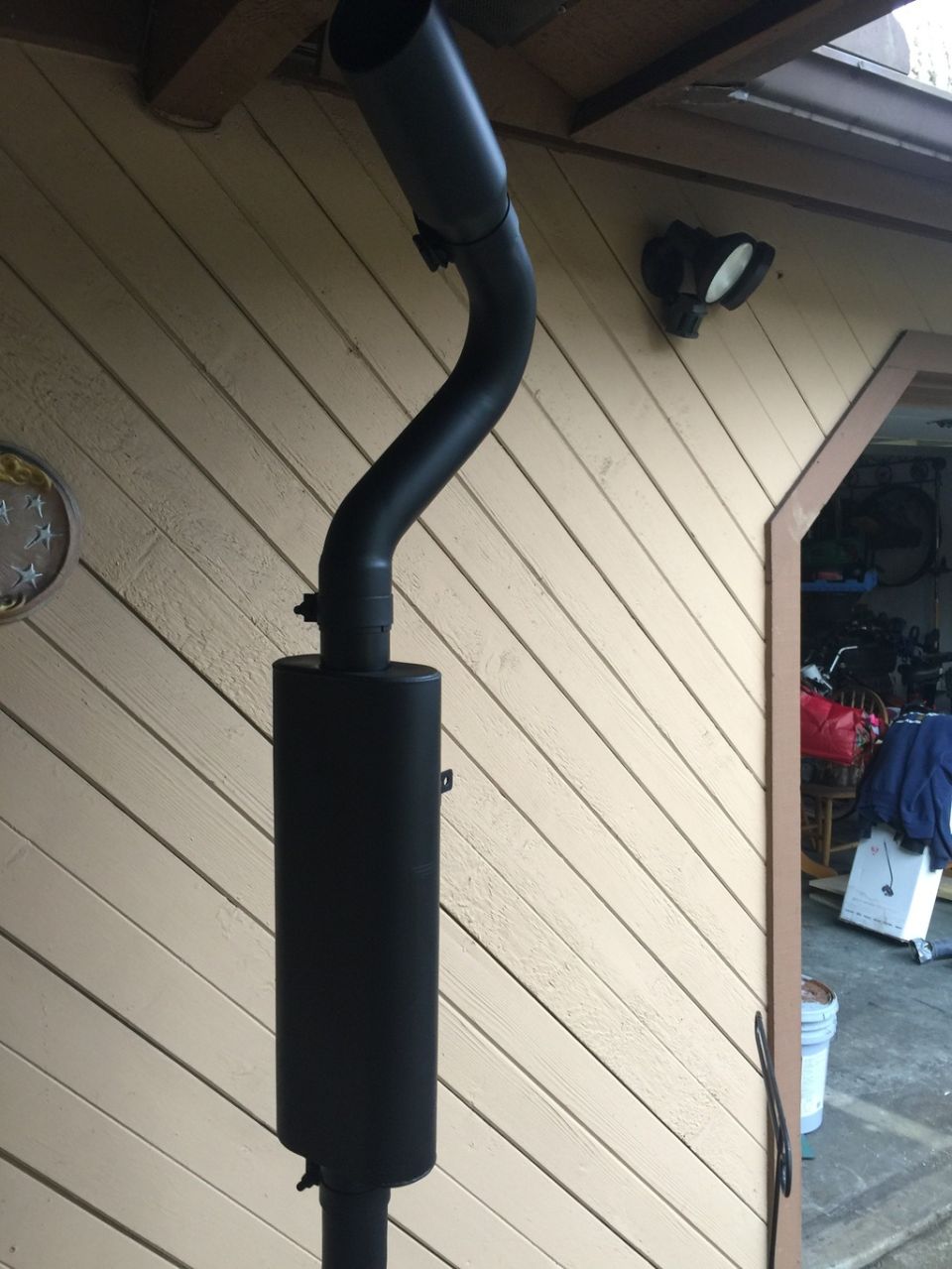 Black exhaust pipe and muffler assembly mounted on a building with tan siding.