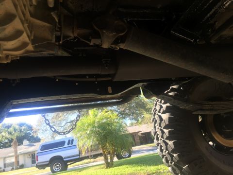 1989 Ford Bronco - The underside of a vehicle, showing the drivetrain components including a driveshaft and exhaust pipe, with a large off-road tire visible o…