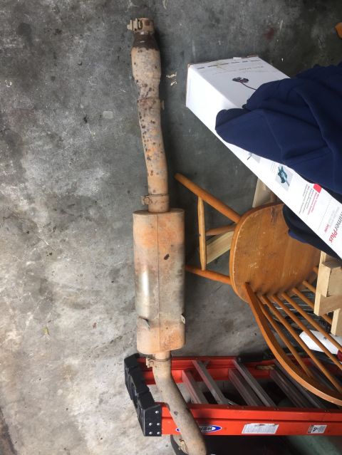 1989 Ford Bronco - A rusty exhaust system, likely from a 1989 Ford Bronco, lies on an orange ladder next to a wooden chair and a cardboard box.